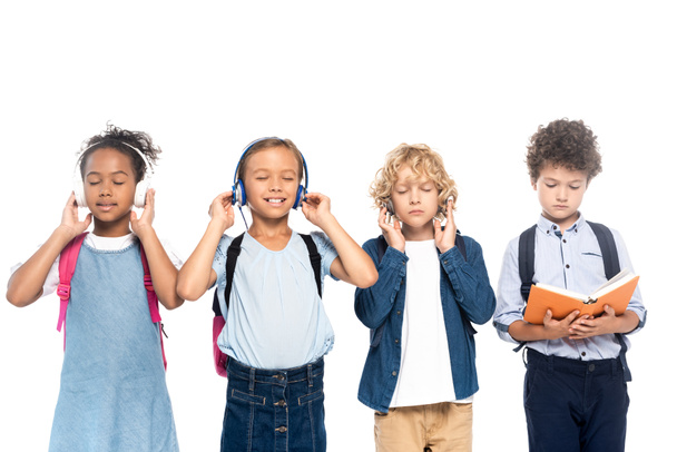 multicultural schoolchildren listening music in wireless headphones near curly boy reading book isolated on white  - Foto, Imagen