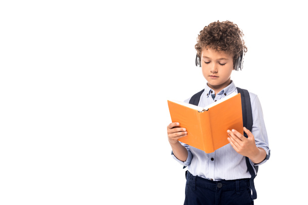 curly schoolboy in wireless headphones reading book isolated on white  - Photo, Image