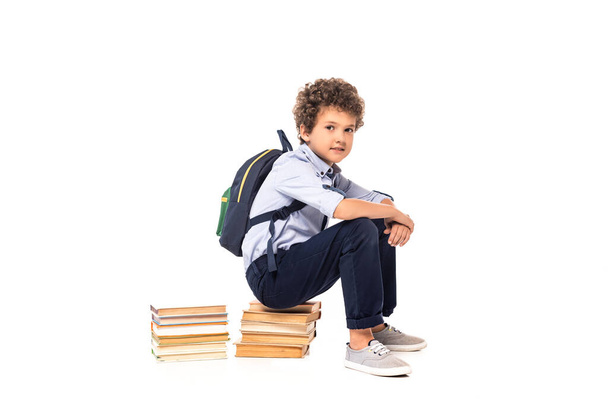 curly schoolboy with backpack sitting on books isolated on white  - Photo, Image