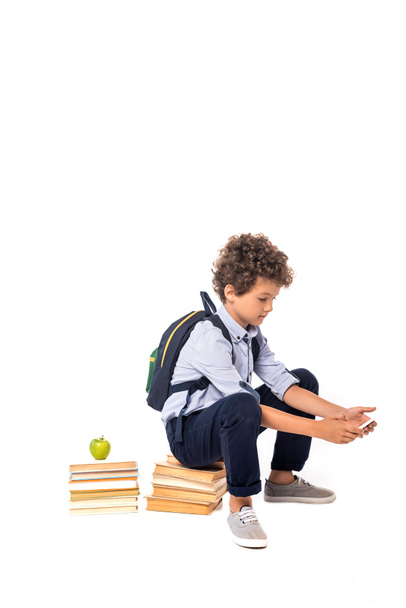 curly schoolboy with backpack sitting on books near apple and using smartphone isolated on white  - Fotoğraf, Görsel