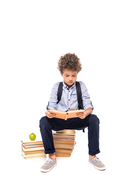 curly schoolboy with backpack sitting on books near apple and reading isolated on white  - Photo, image