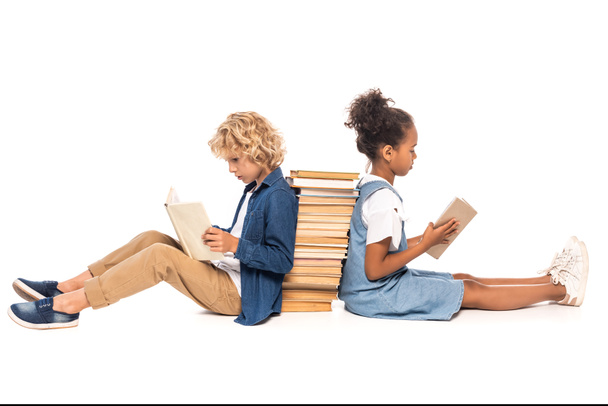 πλαϊνή όψη πολυπολιτισμικών παιδιών που κάθονται κοντά σε βιβλία και διαβάζουν απομονωμένα σε λευκό  - Φωτογραφία, εικόνα