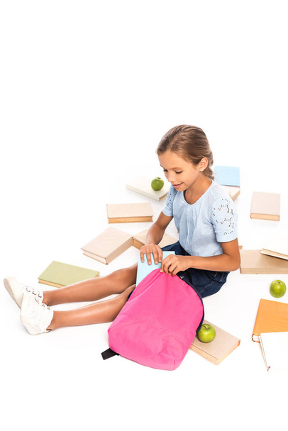 schoolkid sitting near apples and putting book in backpack isolated on white - Photo, Image