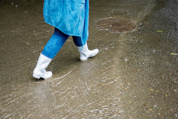 Een vrouw met witte rubberen laarzen loopt door de straat in de regen. Water stroomt langs de weg. Straatscènes in de regen. Regenachtig herfstweer. - Foto, afbeelding