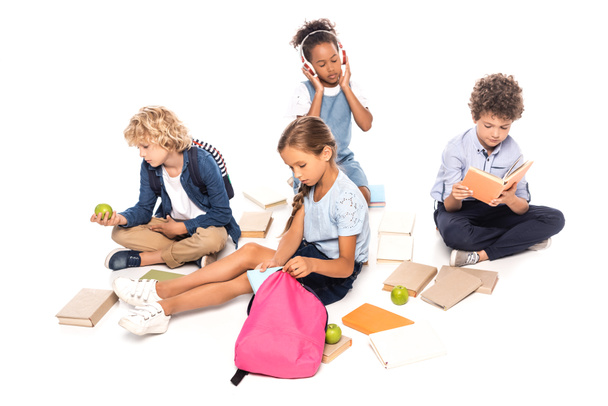 escolares sentados perto de livros, maçãs e criança afro-americana em fones de ouvido sem fio isolados em branco - Foto, Imagem
