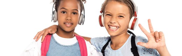 panoramic crop of schoolkid listening music in wireless headphones, hugging african american friend and showing three fingers isolated on white  - Photo, Image