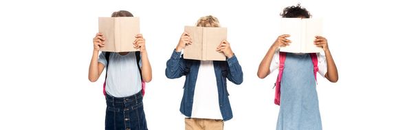 горизонтальное изображение мультикультурных школьниц и школьников, покрывающих лица книгами, изолированными на белом  - Фото, изображение