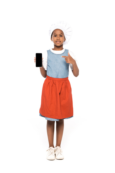 niño afroamericano vestido con traje de chef apuntando con el dedo al teléfono inteligente con pantalla en blanco aislado en blanco  - Foto, Imagen