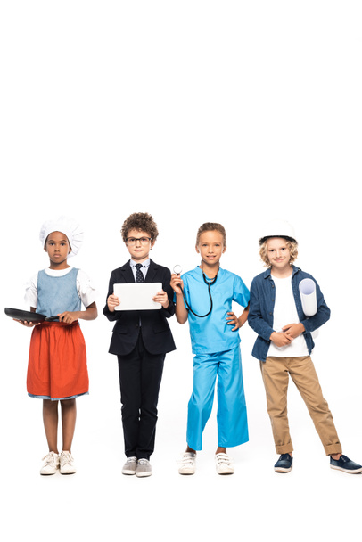 niños multiculturales en trajes de diferentes profesiones que sostienen sartén, plano, estetoscopio y tableta digital aislados en blanco  - Foto, imagen