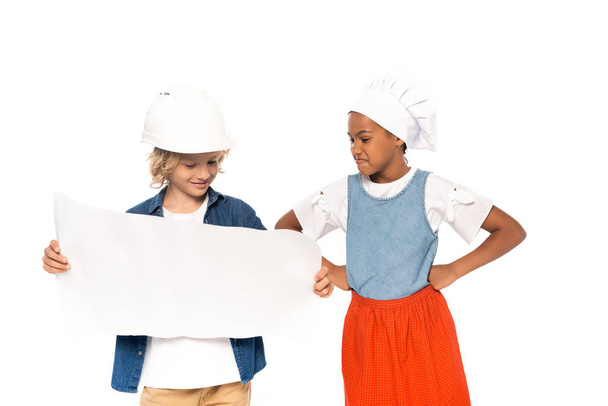 lockiger Junge in Schutzhelm und Architektenkostüm mit Blick auf die Blaupause in der Nähe eines afrikanisch-amerikanischen Kindes mit Kochmütze, die Hände auf Hüften isoliert auf weiß  - Foto, Bild