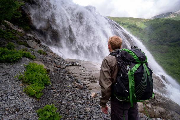 Man hiking with backpack looking at waterfall. Travel Lifestyle adventure concept, active vacations into the wild harmony with nature. North Caucasus, Dombai, Russia. - Foto, Imagem