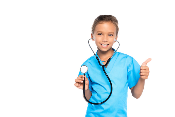 niño disfrazado de médico sosteniendo estetoscopio mientras muestra el pulgar hacia arriba aislado en blanco  - Foto, Imagen
