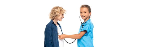 panoramiczne ujęcie dziecka w kostiumie lekarza badającego kręconego chłopca odizolowanego na biało  - Zdjęcie, obraz