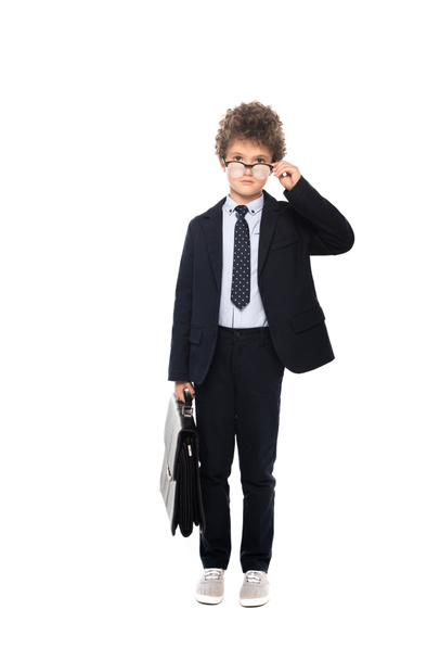 niño rizado en traje tocando gafas y sosteniendo maletín aislado en blanco - Foto, Imagen
