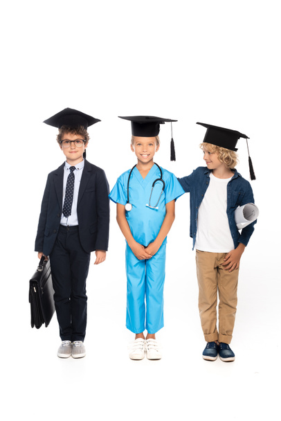 dzieci w czapkach dyplomowych ubrane w kostiumy różnych zawodów stojące z odciętą na biało plandeką, aktówką i stetoskopem  - Zdjęcie, obraz