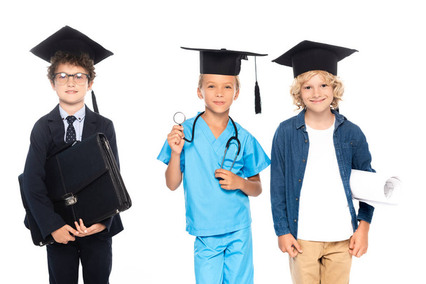 children in graduation caps dressed in costumes of different professions holding blueprint, stethoscope and briefcase isolated on white  - Foto, afbeelding
