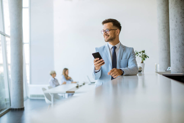 Щасливий молодий чоловік використовує свій мобільний телефон в офісі і посміхається, поки його колеги працюють на задньому плані
 - Фото, зображення
