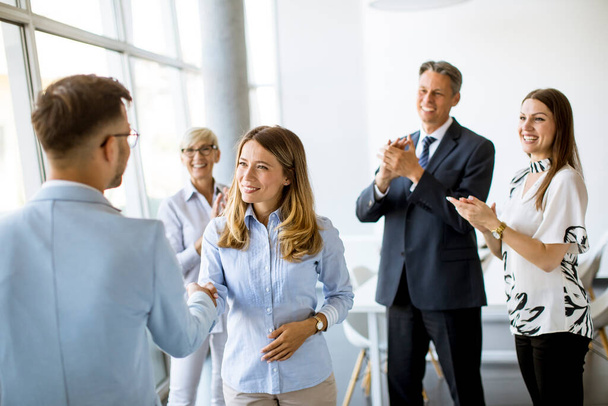 Молодые деловые партнеры пожимают друг другу руки в офисе, пока их команда аплодирует на заднем плане - Фото, изображение