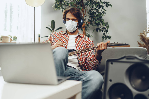 Young man plays guitar at home in medical mask. Coronavirus quarantine lifestyle concept - 写真・画像