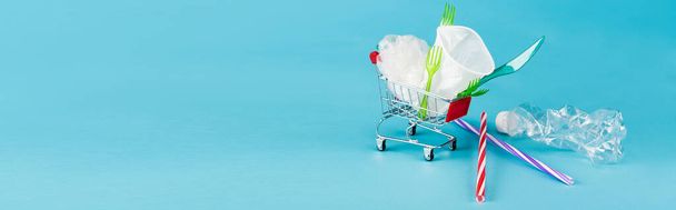 objetos de plástico desechables en un pequeño carrito de compras sobre fondo azul, plano panorámico - Foto, Imagen