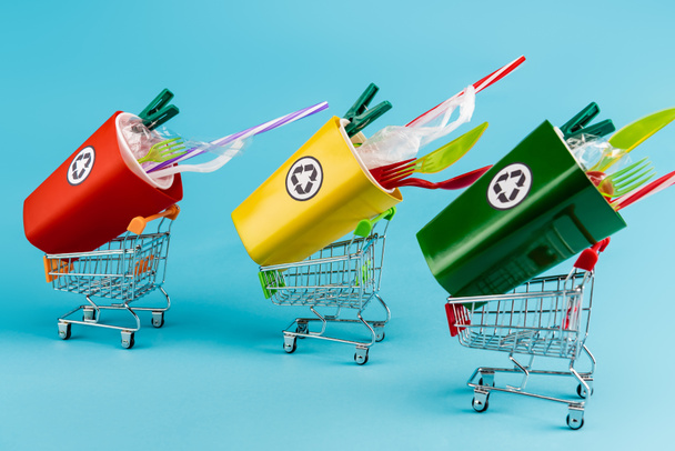 papeleras de reciclaje multicolores con plástico en pequeños carritos de compras sobre fondo azul - Foto, Imagen