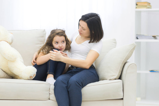 Photo of sad little girl sitting on the sofa and her mom hugging her. - Foto, Imagen