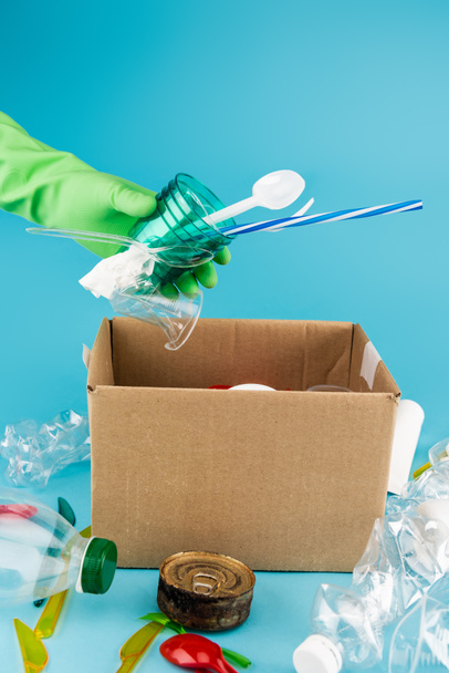vue recadrée du nettoyant dans un gant en caoutchouc ramassant les déchets dans une boîte en carton sur fond bleu - Photo, image