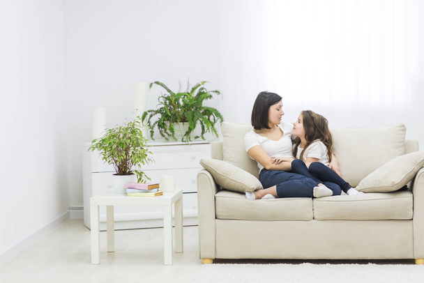 Photo of beautiful mother hugging with her cute little daughter and look each other in living room. - Photo, Image