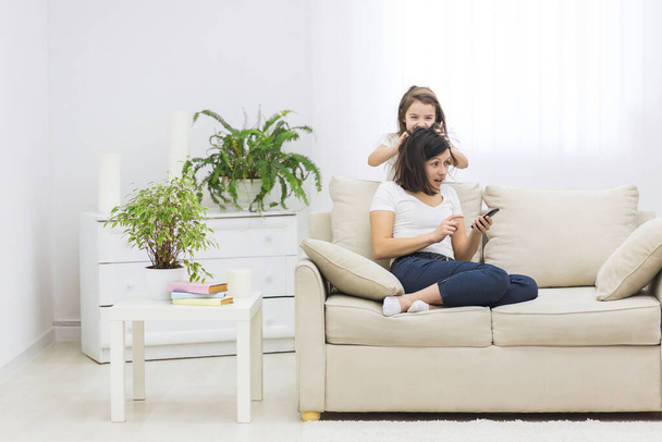Photo of mother sitting on white sofa in her phone and her daughter hiding behind the sofa. - Foto, imagen