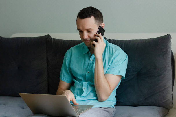 Junger Mann sitzt auf der Couch, telefoniert und arbeitet am Laptop - Foto, Bild