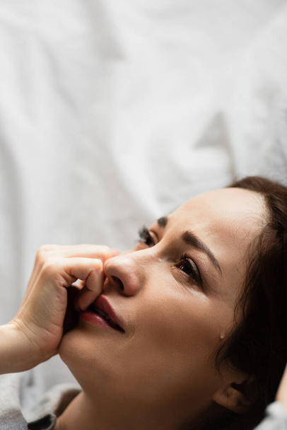 turhautunut ja brunette nainen koskettaa kasvot itkiessään sängyssä  - Valokuva, kuva