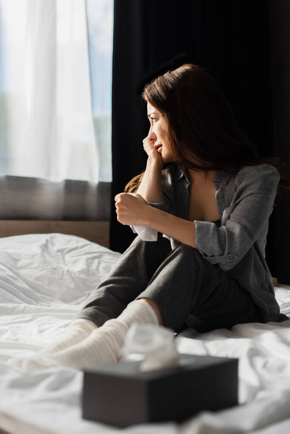 επιλεκτική εστίαση της καταθλιπτικής γυναίκας που κάθεται στο κρεβάτι κοντά σε κουτί ιστού  - Φωτογραφία, εικόνα