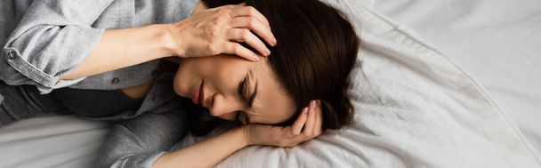 Panoramaaufnahme einer brünetten Frau, die Migräne hat, den Kopf berührt und auf dem Bett liegt - Foto, Bild
