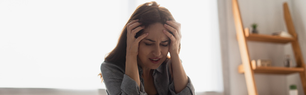 panoramic crop of brunette woman with closed eyes suffering from pain and touching head at home - Photo, Image
