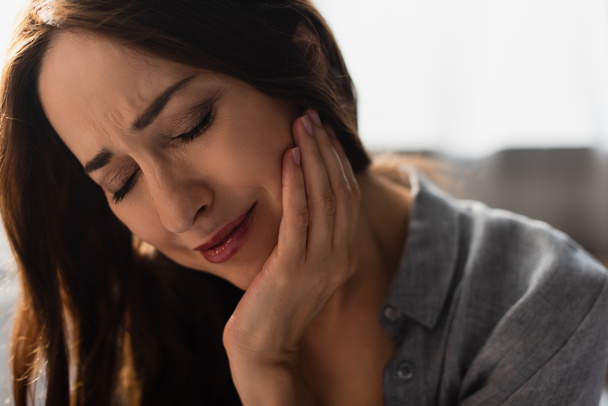 sad brunette woman with closed eyes touching face while suffering from toothache at home - Foto, Bild