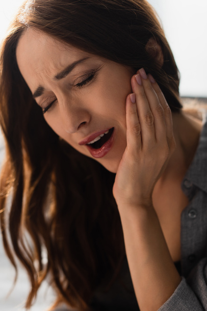 brunette woman with closed eyes and open mouth touching face while suffering from toothache at home - Photo, Image