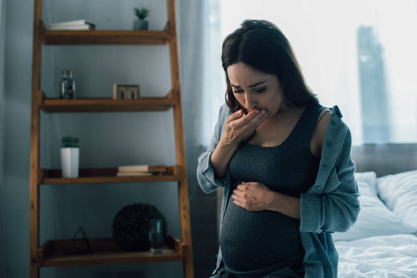 вагітна жінка торкається живота і закриває рот, маючи нудоту вдома
 - Фото, зображення