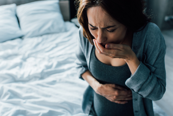 вагітна жінка з закритими очима, що закривають рот, маючи нудоту вдома
 - Фото, зображення