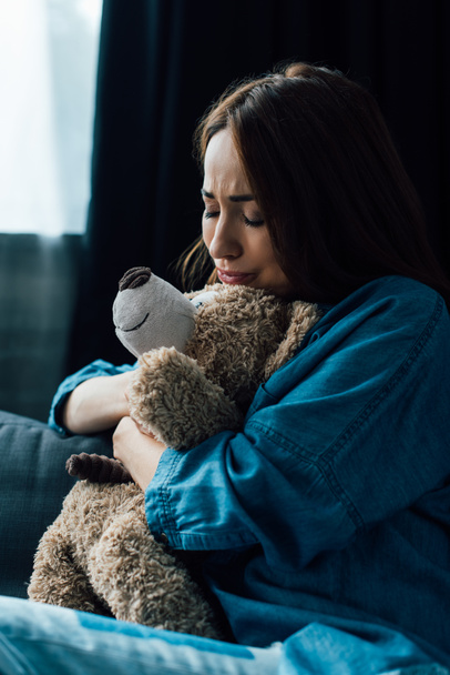 upset brunette woman with closed eyes holding teddy bear in living room  - Photo, Image
