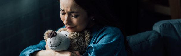 colheita panorâmica de mulher morena triste com olhos fechados segurando ursinho na sala de estar  - Foto, Imagem
