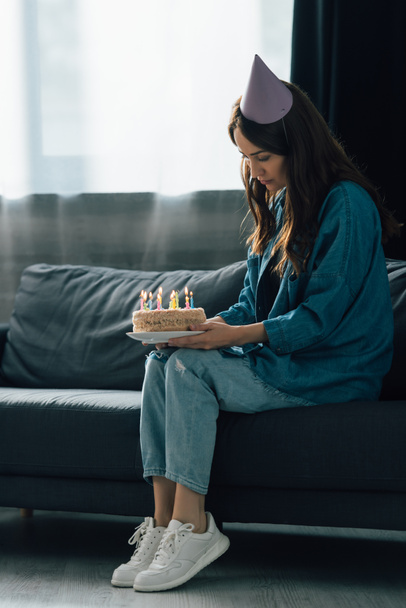sad woman in party cap sitting on sofa and holding birthday cake with candles  - Foto, imagen
