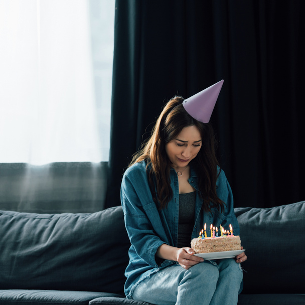 απογοητευμένη γυναίκα με καπέλο κόμμα κάθεται στον καναπέ και κρατώντας τούρτα γενεθλίων με κεριά  - Φωτογραφία, εικόνα
