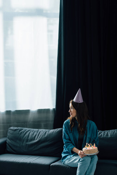 upset woman in party cap sitting on sofa, holding birthday cake with candles and looking away  - Foto, imagen