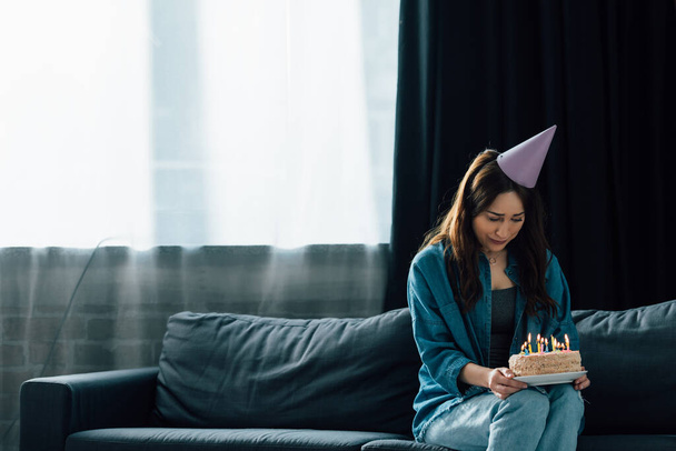 frustrado morena mujer en partido gorra sentado en sofá y celebración de pastel de cumpleaños con velas  - Foto, imagen