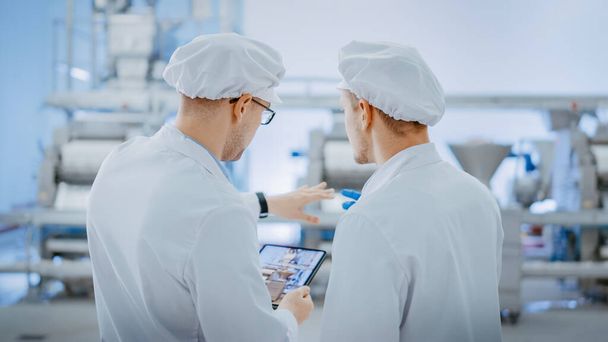Two Young Food Factory Employees Discuss Work-Related Matters. Male Technician or Quality Manager Uses a Tablet Computer for Work. They Wear White Sanitary Hat and Work Robes. - Foto, Bild