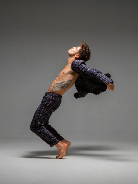 Handsome young guy dancing expressive dance in suit with naked torso isolated on gray background. Body with tattoo - 写真・画像