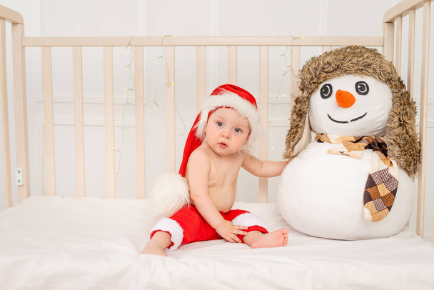 Küçük bir çocuk Noel Baba şapkasıyla beşiğinde oturuyor. Bir kardan adamla. Mutlu yıllar 2021. - Fotoğraf, Görsel