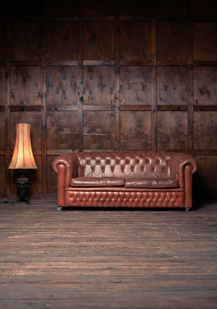 Brown leather sofa in wooden apartment interior against background of wooden wall. Big vintage couch with cozy luminous floor lamp standing next to it. Relax concept. Front view. Vertical format - Foto, imagen