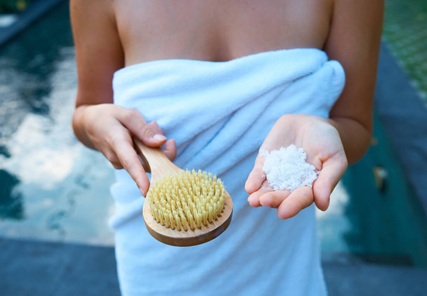 Personal care. A girl in a white towel holds sea salt in her hand and a scrub brush on the background of the pool - Photo, Image