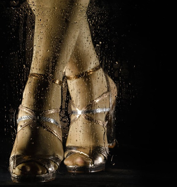 female feet in high heeled sandals behind glass with water drops on black background - Foto, Imagen